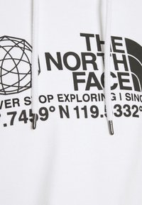 Sweat à capuche blanc avec texte imprimé en noir "THE NORTH FACE" et coordonnées à côté d'un design géométrique de globe et cordons blancs avec embouts métalliques.
