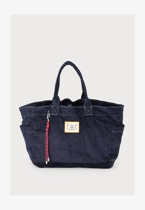 Torba typu tote z sztruksu w kolorze granatowym, z dwoma uchwytami, białymi przeszyciami, logo na froncie i czerwonym plecionym breloczkiem wiszącym na jednym uchwycie.