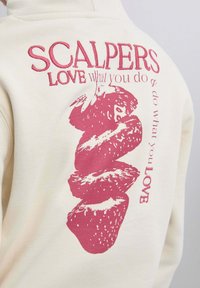 Sudadera crema con bordado rojo de fresas y el texto "SCALPERS LOVE lo que haces" en la parte posterior.