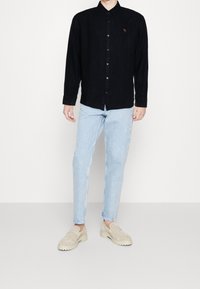 Camisa negra de botones con collar y mangas largas, combinada con jeans de color azul claro y zapatos slip-on beige. La camisa tiene un pequeño detalle de logo.