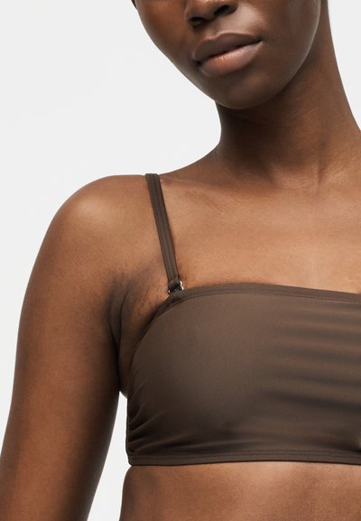 Κοντινό πλάνο ενός ατόμου που φοράει ένα καφέ μπάντεου τοπ με λεπτές ρυθμιζόμενες τιράντες σε απλό ανοιχτό φόντο.