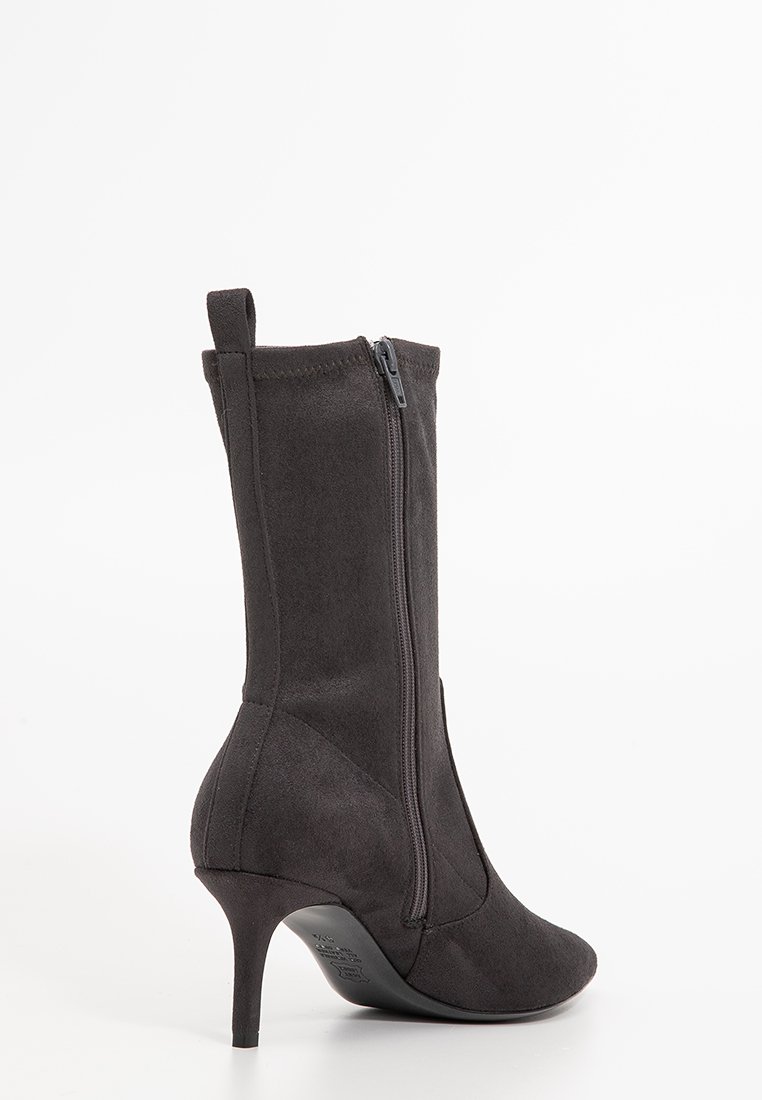 Schwarze Wildleder-Stiefel mit Absatz, bis zur Wade, seitlichem Reißverschluss und Zuglasche, spitzem Schuhkopf und schlankem Stiletto-Absatz auf weißem Hintergrund.
