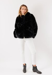 Giacca in pelliccia sintetica nera con un ampio colletto, caratterizzata da un design loose, indossata sopra una maglia nera e pantaloni bianchi, abbinata a stivaletti neri. 