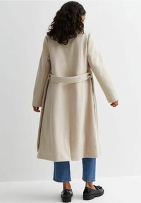 Abrigo de lana beige con cintura ceñida, mangas largas y una abertura en la parte posterior, combinado con jeans azules y zapatos negros. Textura suave, diseño sencillo.