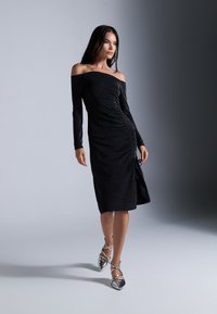 Robe noire à épaules dénudées avec des manches longues, détail froncé sur le côté et éclat métallique ; associée à des talons argentés à brides avec des bouts pointus.
