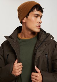 Ciemnobrązowa kurtka puchowa z zamykanymi kieszeniami, w połączeniu z zielonym swetrem o fakturze i brązową dzianinową czapką.