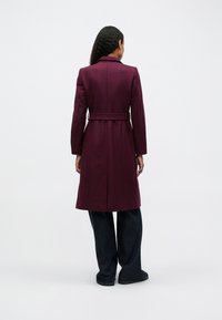 Burgundyröd ullkappa med bälte, långa ärmar och slit på ryggen. Bärs över mörka vida byxor, visad bakifrån mot en enkel bakgrund.