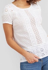 Haut blanc à manches courtes avec broderie à jour, présentant un motif texturé et un ourlet festonné. Tissu lisse avec un col rond.