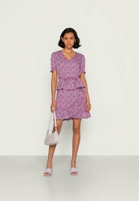 Vestido floral roxo com cintura peplum, mangas curtas bufantes, barra com babados, combinado com sandálias roxas claras e uma pequena bolsa.