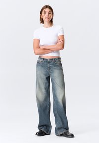 Τζιν με ψηλή μέση και φαρδύ πόδι σε ξεβαμμένο μπλε denim με ελαφρύ ομπρέ εφέ, συνδυασμένο με εφαρμοστό λευκό τοπ με κοντά μανίκια.