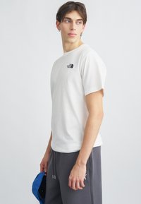 Λευκό βαμβακερό t-shirt με κοντά μανίκια, με μαύρο λογότυπο στο στήθος. Το μοντέλο κρατάει ένα μπλε καπέλο και φοράει γκρι φόρμες.