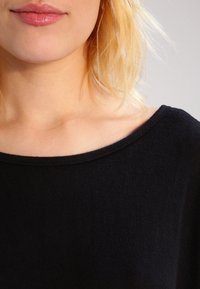 Svart stickad tröja med vid halsringning, mjuk textur och ribbat avslut. Visar nyckelben och nacke, fokuserar på tygets detaljer.