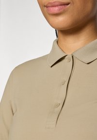 Beige polo-paita, jossa on kaulus, kolme nappia, sileä pintakuvio ja lyhyet hihat, kuvattu kaula-aukosta ylöspäin.