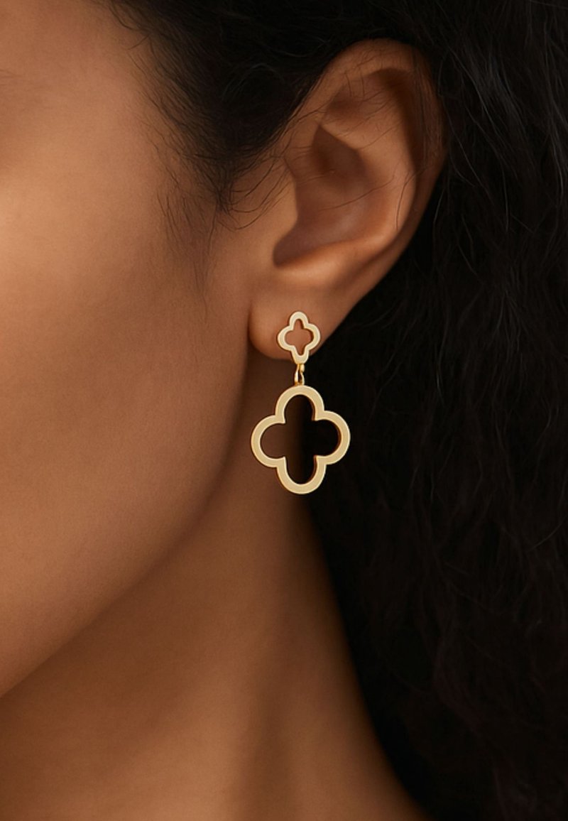 Boucles d’oreilles ton or avec des motifs creux en forme de trèfle portées à l’oreille gauche d’une personne sur fond de cheveux et de peau foncés.