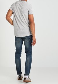 Camiseta gris de manga corta, jeans de mezclilla y zapatos casuales con acentos en contraste. Enfoque en la vista trasera, con costuras visibles y detalles de herrajes.