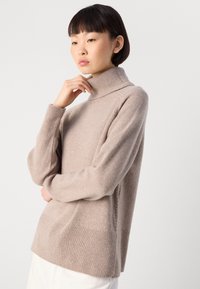 Donna che indossa un maglione a collo alto beige con polsini e orlo a coste, in posa con una mano che tocca il collo e l’altro braccio incrociato.