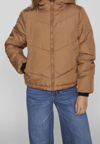 Polstrad, brun jacka med huva och dragkedja. Har horisontell sömnad och elastiska manschetter, tillsammans med vida blå jeans.