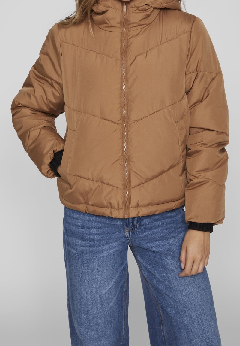 Polstrad, brun jacka med huva och dragkedja. Har horisontell sömnad och elastiska manschetter, tillsammans med vida blå jeans.