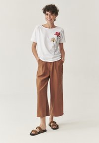 T-shirt blanc à manches courtes avec imprimé floral ; pantalon large marron ; sandales marron avec accents dorés. Fond uni, coupe décontractée.