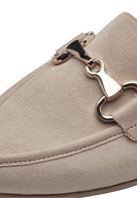 Mocassin en daim beige avec détail en métal doré en forme de mors de cheval et coutures visibles sur le dessus et les bords latéraux.