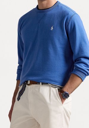 Mand iført en blå langærmet sweatshirt med et lille broderet logo, hvide bukser, stribet bælte og et armbåndsur med blå urskive.