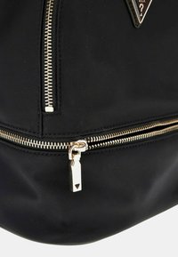 Zaino in nylon nero con zip e hardware dorati, caratterizzato da un accento triangolare con il logo del brand sul davanti. Texture liscia con design strutturato.