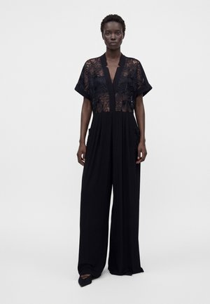 Femme portant une combinaison noire avec un haut en dentelle florale transparente et un pantalon à jambes larges, debout devant un fond clair uni.