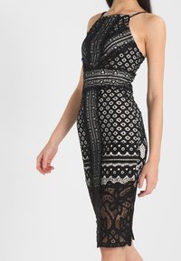 Robe en dentelle noire présentant des motifs géométriques complexes, sans manches, et une silhouette ajustée. L'ourlet se termine juste en dessous du genou.