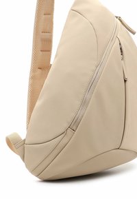 Beige ryggsäck tillverkad av lätt, smidigt material. Har ett strukturerat bälte, vinklad form och en sidficka med dragkedja för förvaring.