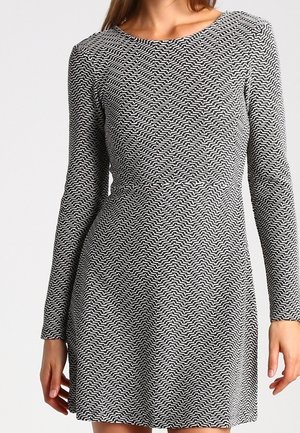 Zwart-witte jurk met lange mouwen, voorzien van een golvend lijnpatroon, een ronde halslijn en een aangesloten lijfje dat overgaat in een uitlopende zoom.