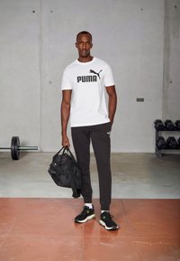 White short-sleeve t-shirt with black Puma logo, black sweatpants, black sneakers with green accents, and a black gym bag.