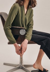 Blusa verde oliva con scollo a V, dettagli con bottoni e maniche lunghe. Modella seduta su una sedia di legno, indossa jeans scuri e sandali neri.