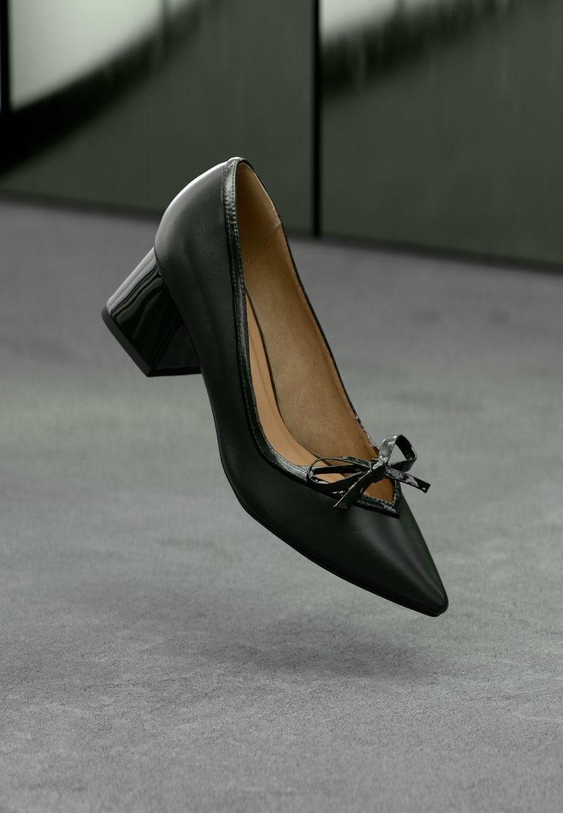Sandal de couro preto com bico apontado, salto bloco e um pequeno laço decorativo, sobre um piso cinza, com parede verde escura ao fundo.