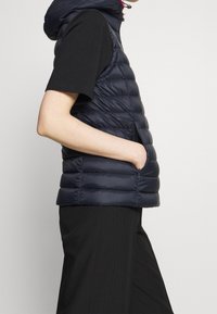 Granatowa kamizelka puchowa z kapturem, z poziomymi pikowanymi sekcjami, zapinaną na zamek boczną kieszenią oraz lekką, błyszczącą fakturą.