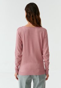 Donna con lunghi capelli castani che indossa un maglione rosa a maniche lunghe e pantaloni grigi chiari, in piedi di fronte a uno sfondo bianco semplice.