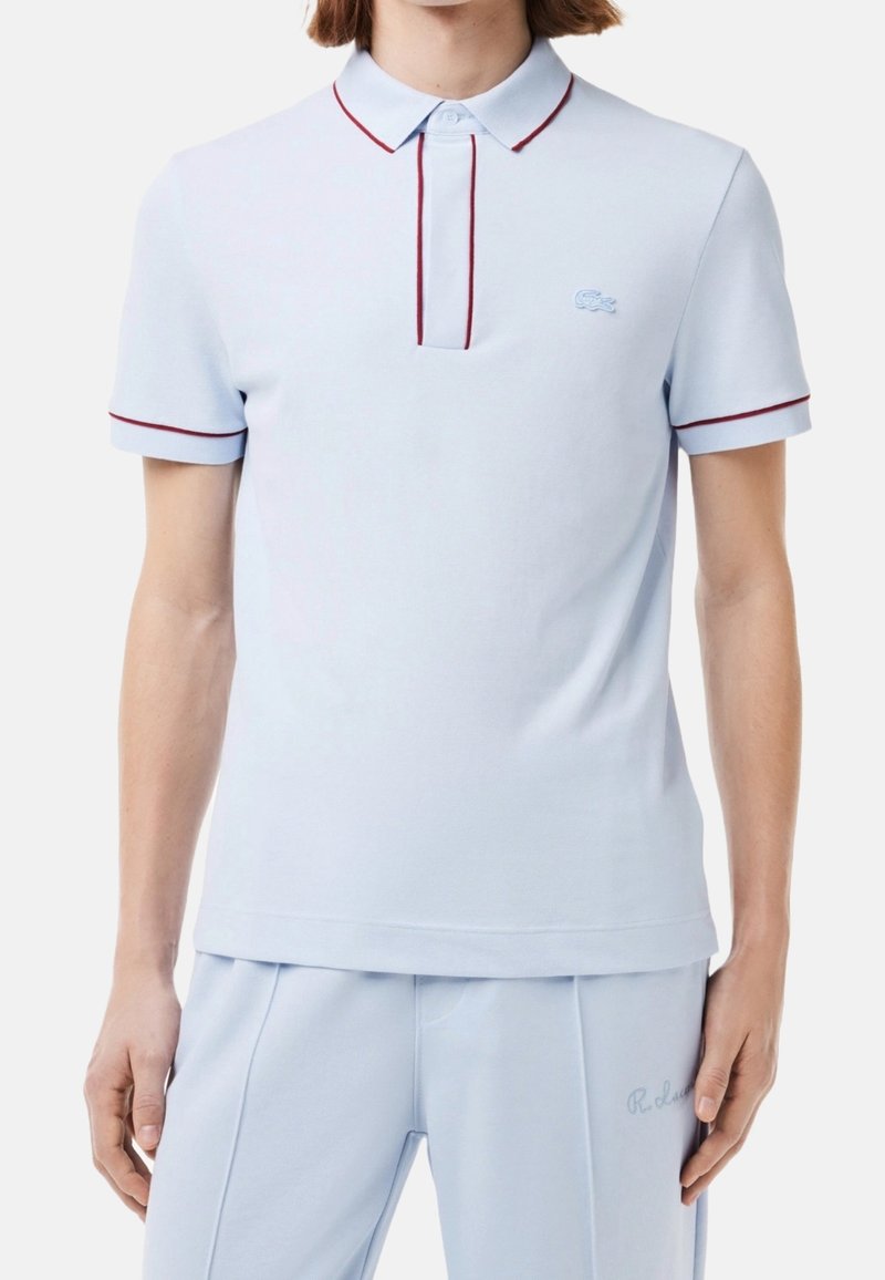 Polo shirt bleu clair à manches courtes avec des bordures bordeaux sur le col et les manches, porté avec un pantalon bleu clair assorti avec des détails brodés.