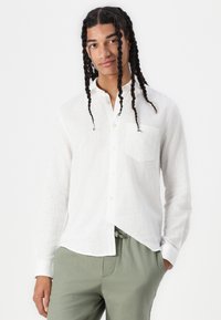 Chemise en lin blanche à manches longues avec un ourlet incurvé, poche avant et boutons sur la patte de boutonnage, assortie à un pantalon vert clair.