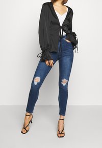 Blouse en satin noir à manches dramatiques nouées aux poignets, associée à un jean skinny bleu taille haute avec des genoux déchirés et des talons à lanières.