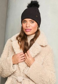 Căciulă neagră din tricot cu pom-pom în vârf și un accent decorativ de culoare aurie; modelul poartă o haină bej din blană sintetică cu un guler larg.