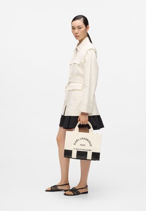 Femme en veste crème ceinturée et jupe noire tenant un sac cabas Karl Lagerfeld blanc et noir, portant des sandales noires à brides, sur fond blanc.