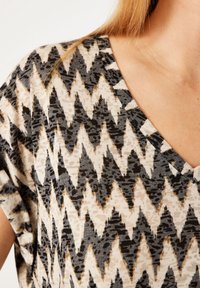 Blusa de cuello en V en una tela ligera con un patrón de zigzag en tonos negro, beige y marrón, con mangas cortas y sueltas.