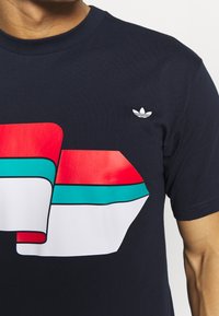 Tmavě modré bavlněné tričko s velkým geometrickým motivem v červené, tyrkysové a bílé barvě, s malým bílým logem Adidas na hrudi.