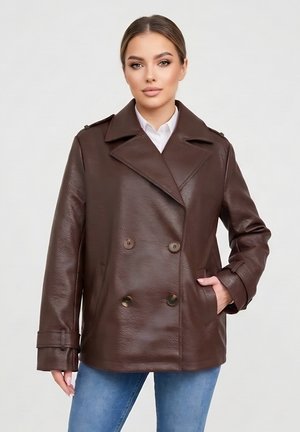 Vrouw met een donkerbruine dubbelgeknoopte leren jas met brede revers en blauwe spijkerbroek, staand tegen een effen lichte achtergrond.