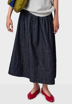 Donna che indossa una gonna plissettata blu navy, un top grigio, porta una giacca verde e scarpe ballerine rosse mentre sta di fronte a uno sfondo neutro.