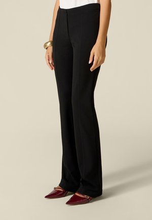 Frau trägt schwarze, maßgeschneiderte Hose, weißes Top, goldenes Armband und bordeauxrote spitze Schuhe und steht vor einem schlichten Hintergrund.