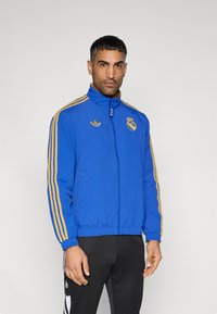 Royal blue track jacket with gold stripes, high collar, zipper front, featuring the Real Madrid logo and Adidas logo on the chest.