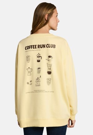 Donna che indossa una felpa giallo pallido con illustrazioni a tema caffè e il testo "COFFEE RUN CLUB" sul retro.