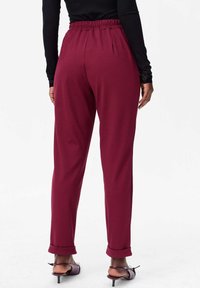 Pantalons bordeaux extensibles avec une taille élastique, jambes effilées et ourlets roulés, associés à un haut sombre à manches longues et des chaussures à talons.