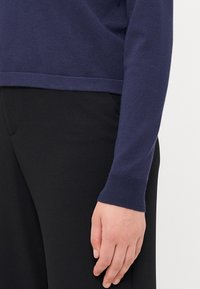 Mørkeblå strikkegenser med ribbestrikkede mansjetter, vist med svarte bukser. Myk tekstur og tettsittende design med rett nederkant.