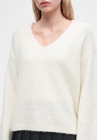Jeune femme portant un pull doux blanc à col en V et une jupe noire, montrée des épaules à la taille sur un fond uni.
